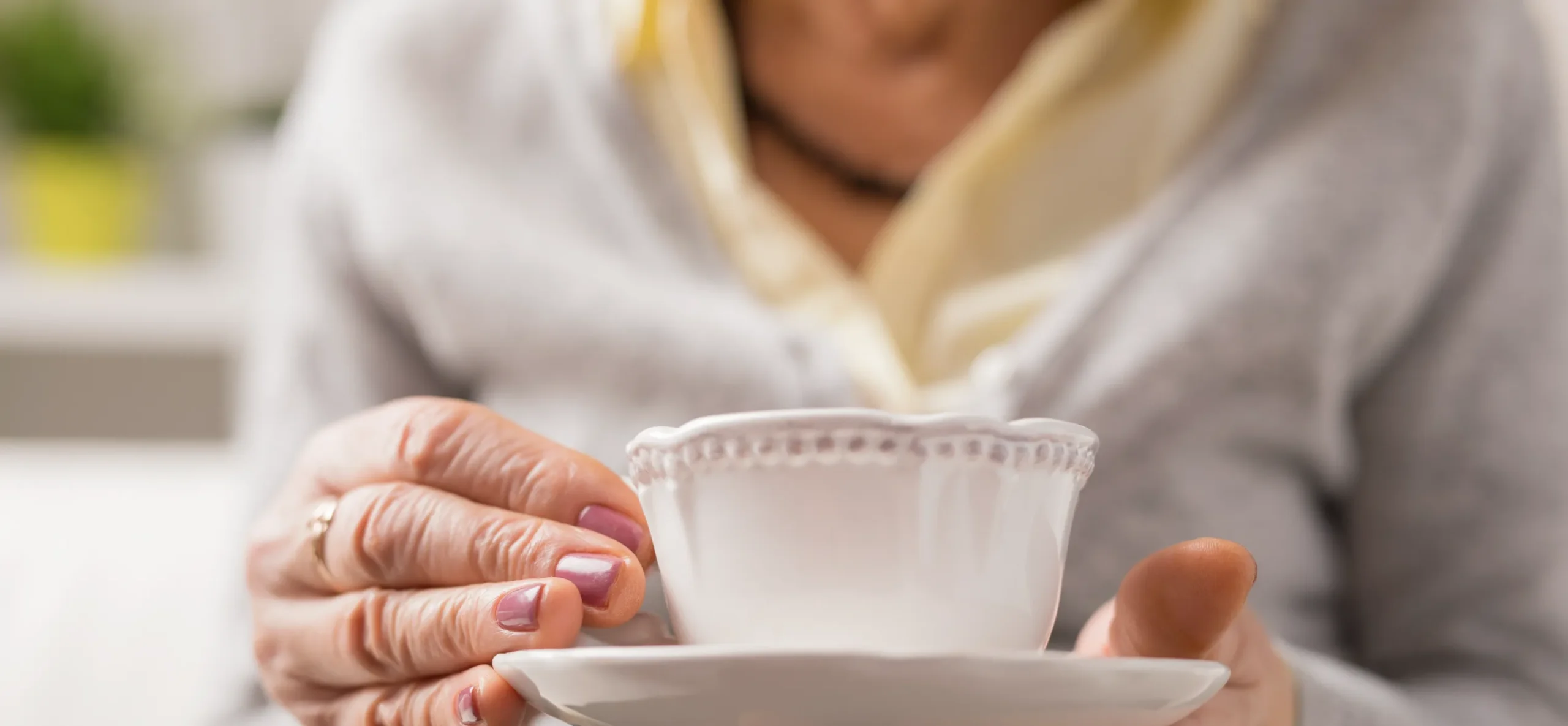 Older woman holding a white teacup and saucer, wearing a gray sweater and light yellow blouse.