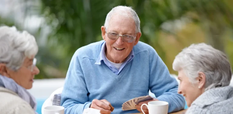 Three older adults sit at a table outside, drinking coffee and playing cards, smiling and talking together.