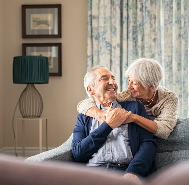 Smiling elderly couple sitting on a couch, hugging and looking at each other in a cozy living room.