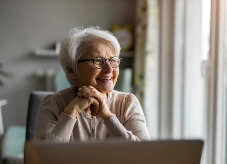 Smiling elderly woman with glasses sits indoors, hands clasped, looking out a window with soft sunlight.