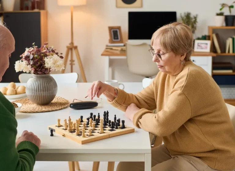 Two older adults sit at a table playing chess in a cozy, well-lit room with flowers and home decor in the background.