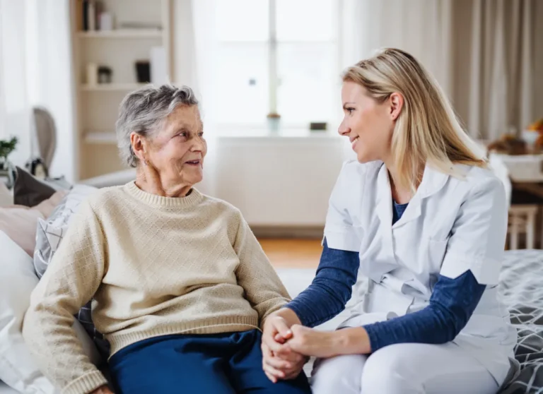 A nurse in white uniform sits and holds hands with an elderly woman, both smiling warmly at each other in a bright room.