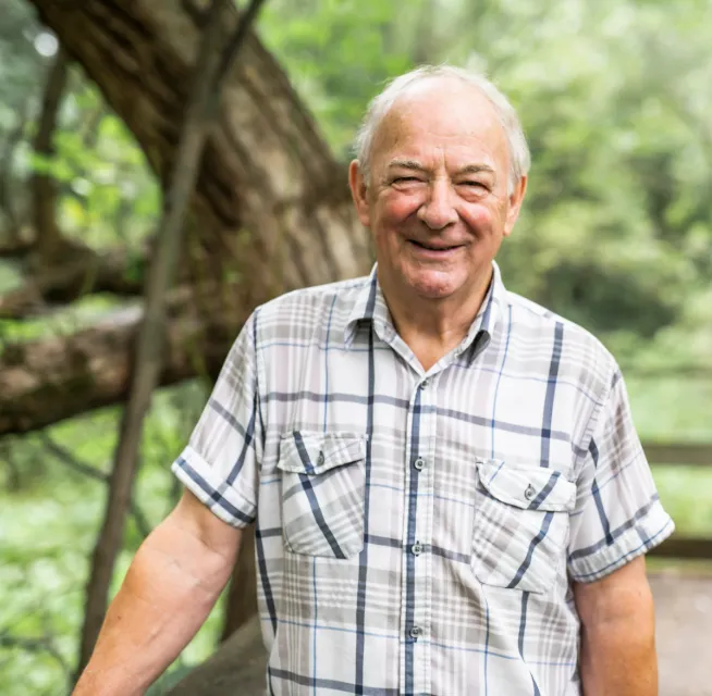 Smiling elderly man in a plaid shirt standing outdoors in a lush, green, wooded area.