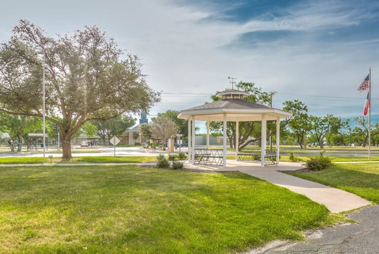 A white gazebo stands on green grass near trees, with flags and a road in the background under a partly cloudy sky.